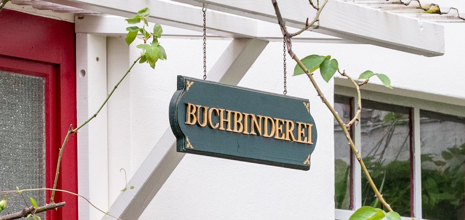 Grünes BUCHBINDEREI-Schild hängt unter Holzbalken.