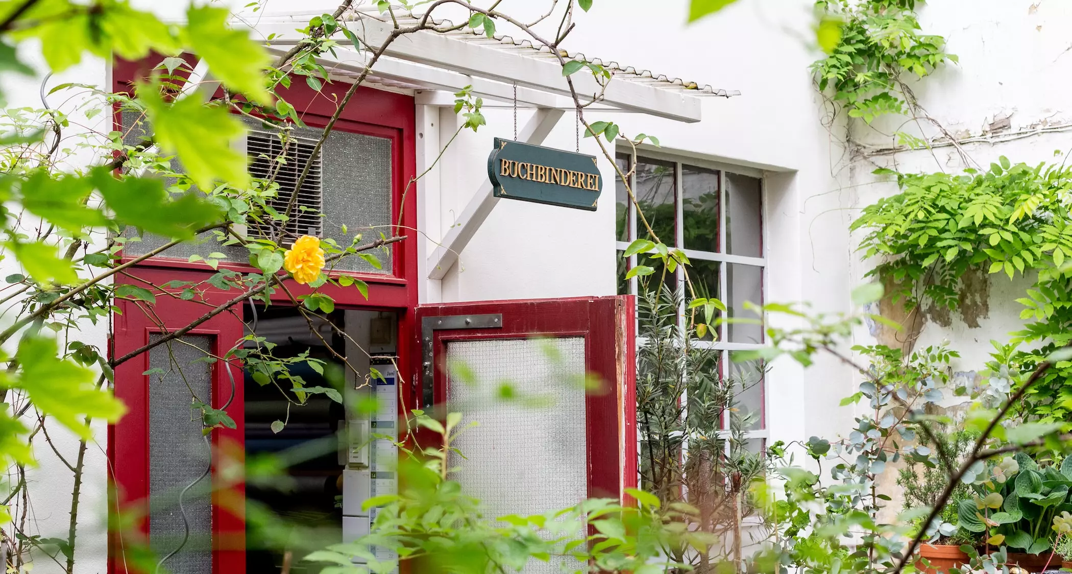 BUCHBINDEREI-Schild über roter Tür an weißem Gebäude mit Pflanzen.